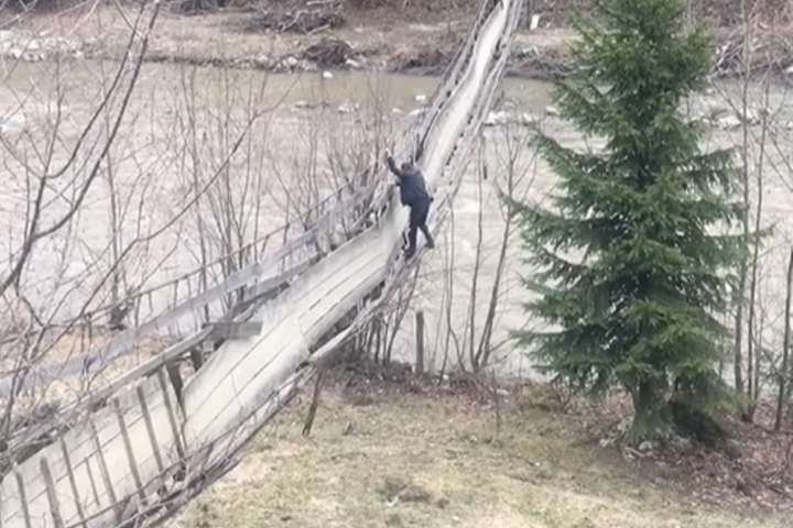 Чоловік, тримаючись за опору вгорі, намагається пройти мостом на інший берег річки